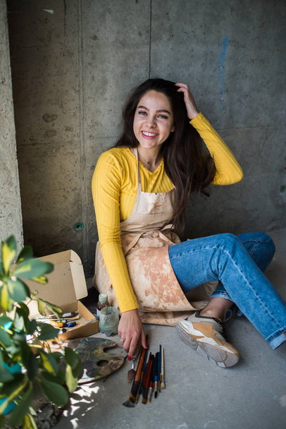 Jonge mooie dame artiest in schort met verf vlekken zitten op de vloer in haar studio - Foto, afbeelding