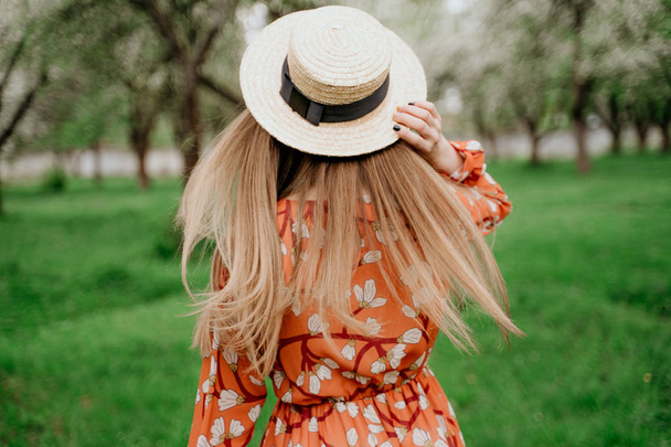 Young beautiful blonde woman in blooming garden. Spring trees in bloom. Orange dress and straw hat. - Valokuva, kuva