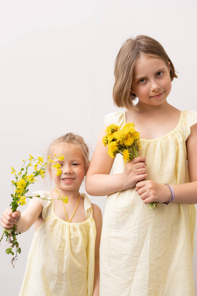 dvě sestry stojí se žlutými květy ve žlutých šatech u bílé zdi - Fotografie, Obrázek