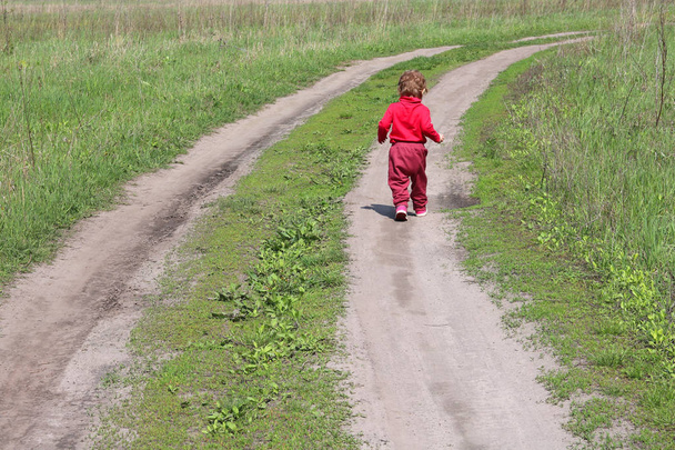 赤い服を着た小さな子供を背中から持つ抽象的な写真は、成長する子供の象徴として緑の草の間の道路に沿って遠くに走り去り、子供たちが大人になりたいという願望 - 写真・画像