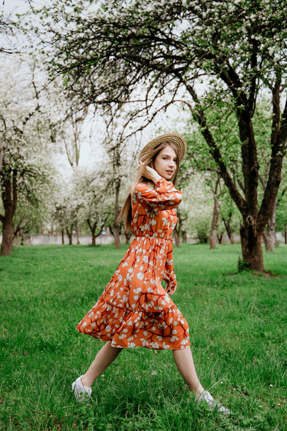 Young beautiful blonde woman in blooming garden. Spring trees in bloom. Orange dress and straw hat. - Fotografie, Obrázek