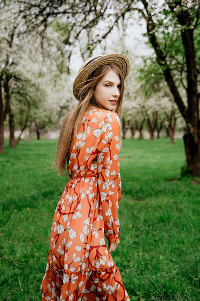 Young beautiful blonde woman in blooming garden. Spring trees in bloom. Orange dress and straw hat. - Foto, Bild