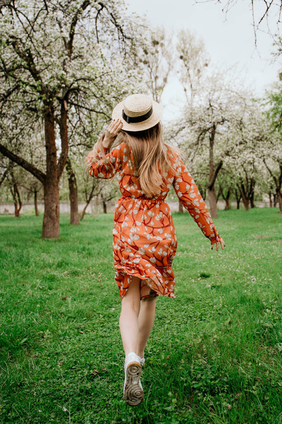Young beautiful blonde woman in blooming garden. Spring trees in bloom. Orange dress and straw hat. - Фото, изображение