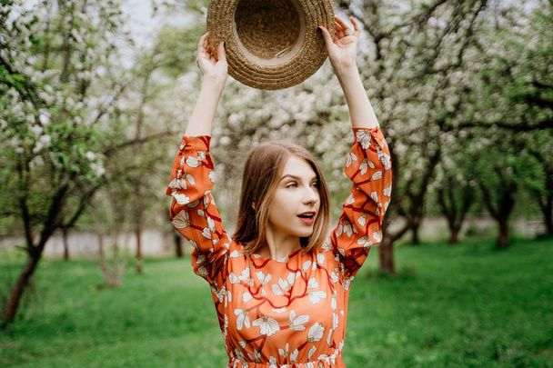 Young beautiful blonde woman in blooming garden. Spring trees in bloom. Orange dress and straw hat. - Foto, Imagem