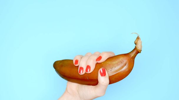 female hands with red nails hold a red banana on a blue background - Foto, Imagen
