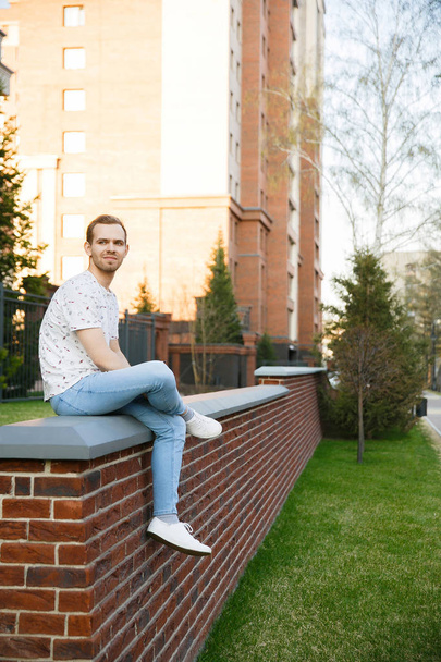 Young beautiful stylish man with bristles sitting on a brick fence near the building on a summer evening - Фото, изображение