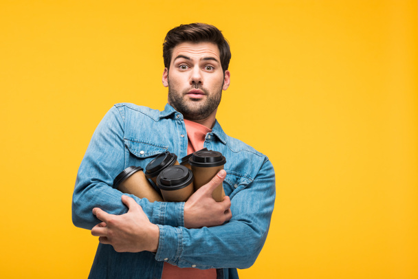 handsome shocked man holding paper cups with coffee to go isolated on yellow - Photo, Image