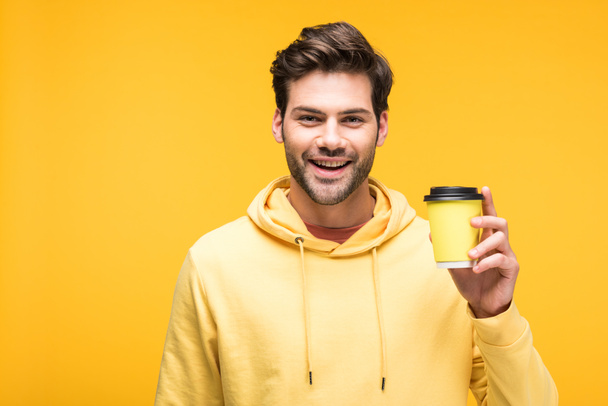 handsome man in hoodie holding coffee to go isolated on yellow - Fotó, kép