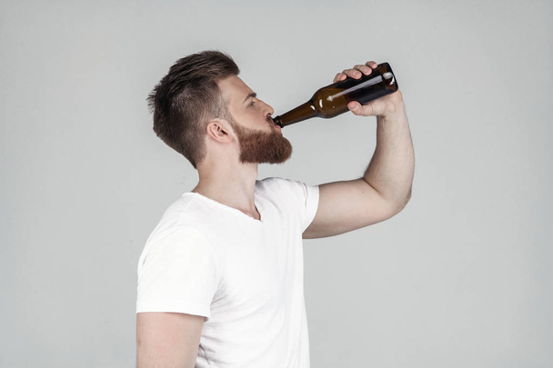Πορτρέτο ενός όμορφου σέξι άντρα με μούσι ντυμένο με λευκό μπλουζάκι που στέκεται στο προφίλ και πίνει μπύρα, στέκεται μπροστά από ένα λευκό φόντο - Φωτογραφία, εικόνα