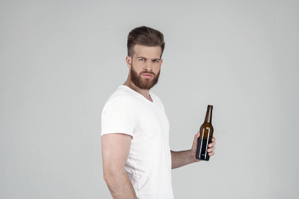 Een portret van een mooie sexy bebaarde man gekleed in een wit T-shirt staat in profiel en drinkt bier en kijkt naar de camera. Hij staat voor de witte achtergrond - Foto, afbeelding