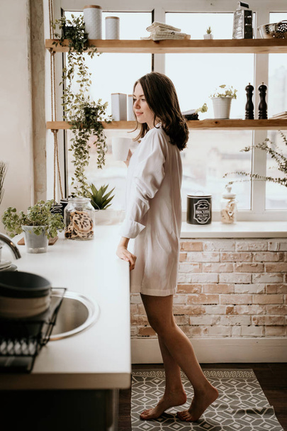 Successful and confident woman on a morning coffee break, resting and enjoying the drink. Young girl in white panties and long shirt with naked shoulder and chest stand on the kitchen of the house - Фото, зображення