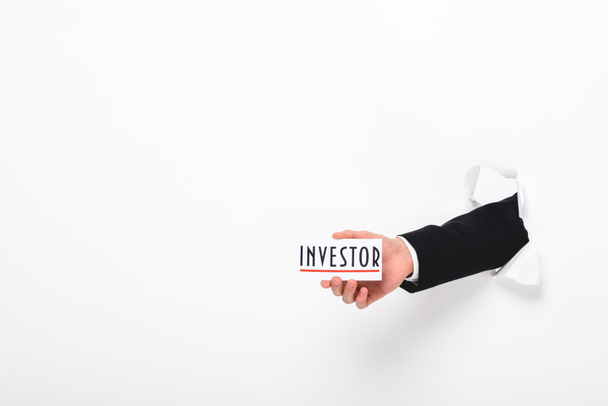 Обрезанный вид человека, держащего карту со словом инвестора через отверстие в бумажной стене на белом
 - Фото, изображение