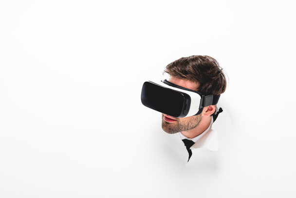 bearded man in Virtual reality headset behind hole in wall on white with copy space - Foto, afbeelding