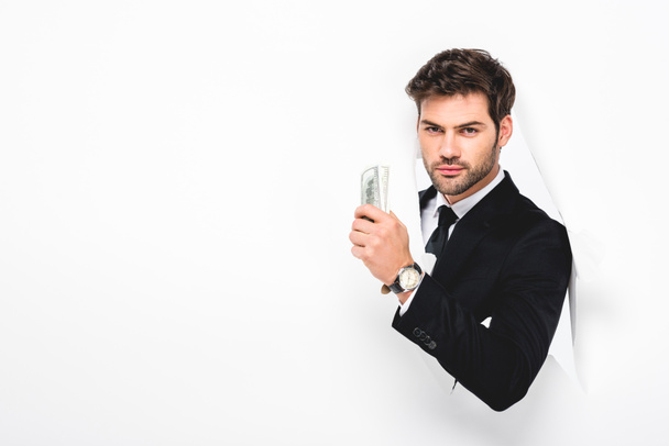 handsome businessman holding dollar banknotes behind hole in paper wall on white - Фото, изображение