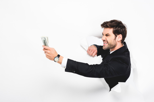 hombre de negocios enojado sosteniendo billetes de dólar detrás de un agujero en la pared de papel en blanco
 - Foto, Imagen
