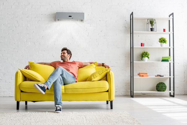 hymyilevä komea mies istuu keltaisella sohvalla ilmastointilaitteen kotona
 - Valokuva, kuva