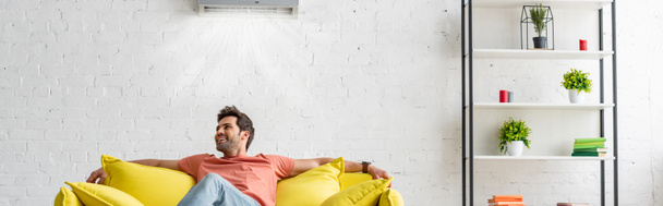 panoramic shot of handsome man sitting on yellow sofa under air conditioner at home - 写真・画像
