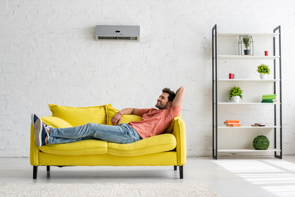 knappe lachende man liggend op gele sofa onder airco thuis - Foto, afbeelding
