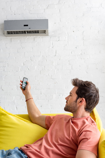 handsome young man holding remote control while lying on sofa under air conditioner - Foto, Imagem
