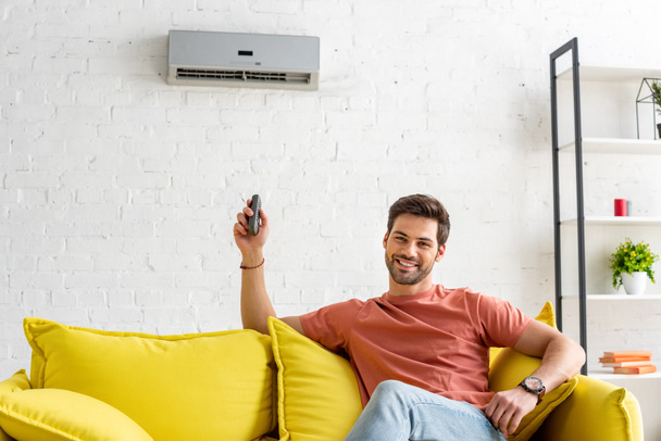 handsome man looking at camera while sitting under air conditioner and holding remote control - Foto, Imagem