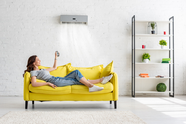 attractive young girl lying on yellow sofa under air conditioner at home - Фото, изображение