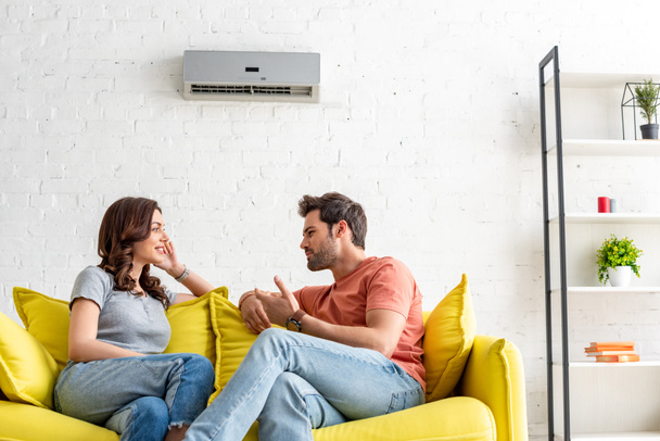 cheerful man and woman talking while sitting on yellow sofa under air conditioner at home - Foto, Imagem