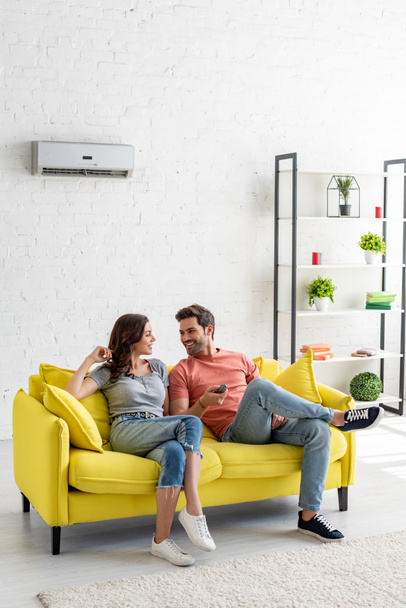 young man and woman talking while sitting on yellow sofa under air conditioner at home - Foto, Imagen