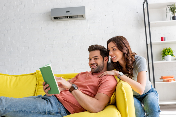 handsome man reading book while lying on sofa under air conditioner near smiling woman - Фото, изображение