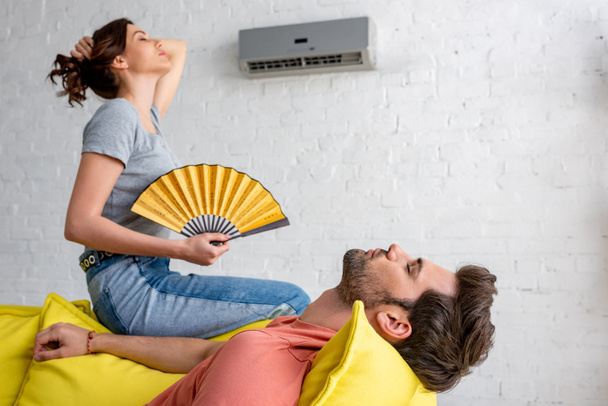 young woman with hand fan sitting on yellow sofa under air conditioner near lying man  - Photo, Image