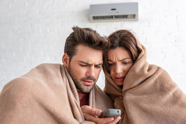 frozen man and woman warming under blankets while sitting under air conditioner with remote control - Photo, Image