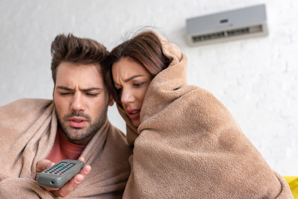 κατεψυγμένο ζευγάρι θέρμανση κάτω από κουβέρτες, ενώ κάθονται κάτω από το κλιματιστικό με τηλεχειρισμό - Φωτογραφία, εικόνα