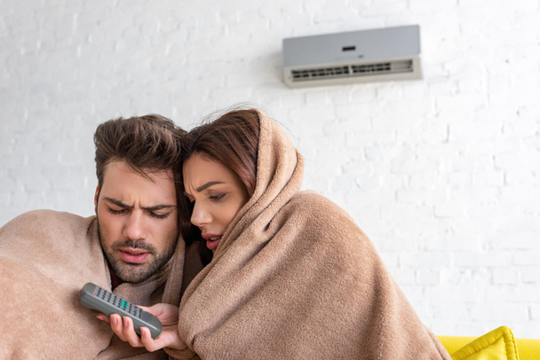 pretty woman holding remote control while warming under blanket with handsome boyfriend - Foto, Imagem