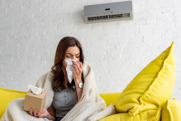mulher doente com guardanapo de papel aquecendo sob cobertor enquanto sentado sob ar condicionado em casa
 - Foto, Imagem