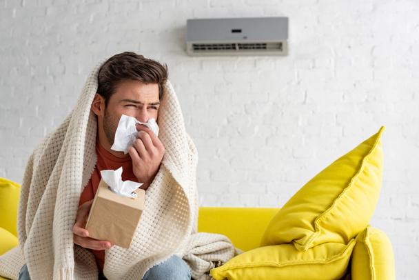 hombre enfermo con servilletas de papel calentándose debajo de la manta mientras está sentado bajo el aire acondicionado en casa
 - Foto, Imagen