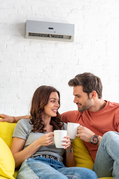 счастливая улыбающаяся пара держит чашки, сидя под кондиционером дома
 - Фото, изображение