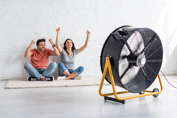 excited man and woman sitting on floor by white wall in front of blowing electric fan - Photo, Image