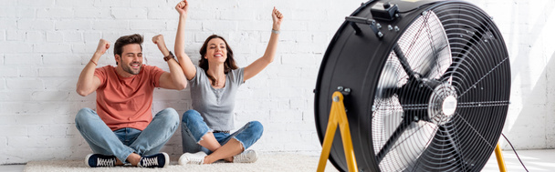 panoramic shot of excited man and woman sitting on floor with raised hands in front of blowing electric fan - Photo, Image