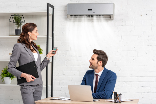 Lächelnde Geschäftsfrau mit Fernbedienung, während sie neben einem gutaussehenden Manager steht, der am Arbeitsplatz unter einer Klimaanlage sitzt - Foto, Bild
