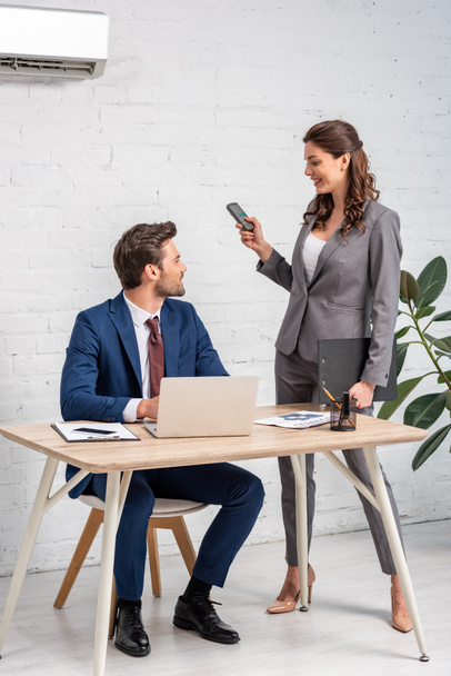 attraktive Geschäftsfrau mit Fernbedienung, während sie in der Nähe eines gutaussehenden Mannes mit Laptop am Arbeitsplatz steht - Foto, Bild