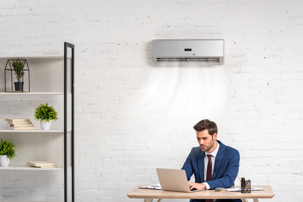 komea liikemies käyttää kannettavaa tietokonetta istuessaan työpaikalla ilmastointilaitteessa
 - Valokuva, kuva