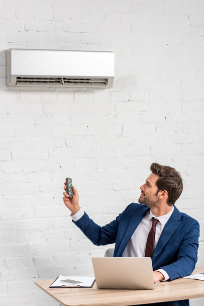 handsome businessman holding remote control while sitting at workplace under air conditioner - Fotoğraf, Görsel