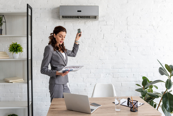 beautiful businesswoman reading business newspaper while standing under air conditioner with remote control - Foto, Imagen