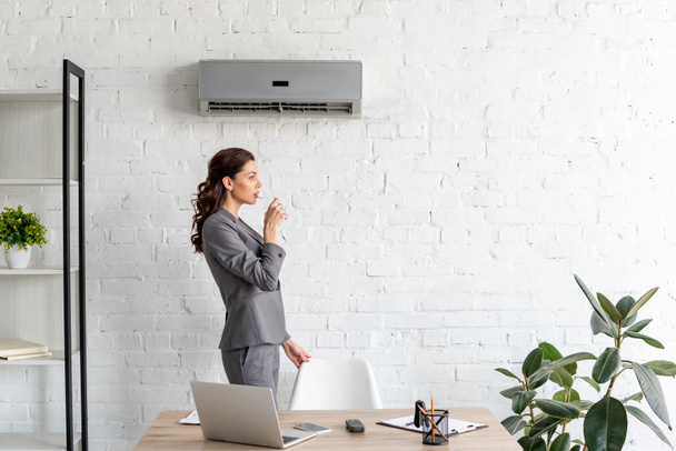 pretty businesswoman drinking water while standing near workplace under air conditioner - Photo, Image