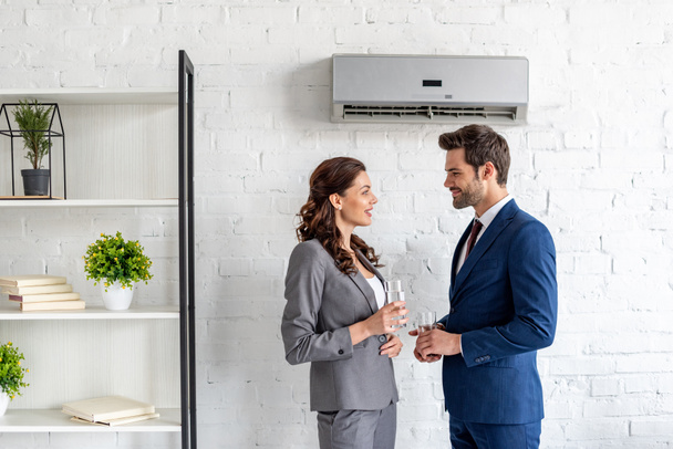 young smiling businesspeople talking while standing under air conditioner with glasses of water - Photo, Image
