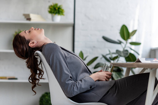 mujer de negocios agotada sentada en la silla con los ojos cerrados y sosteniendo un vaso de agua mientras sufre de calor en la oficina
 - Foto, imagen