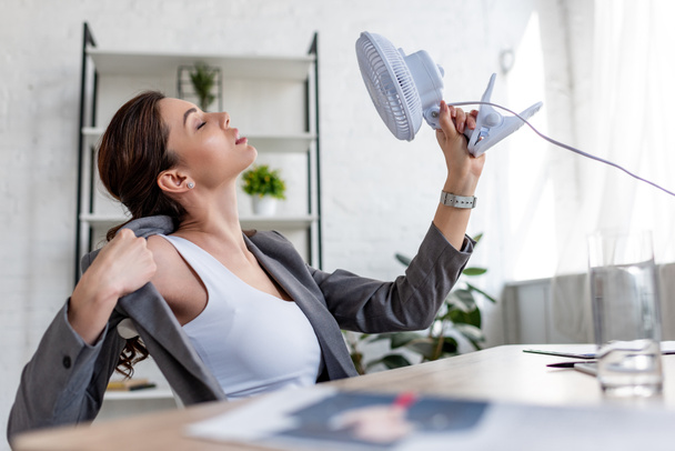 selektivní zaměření krásné obchodní ženy na teplo a držící elektrický ventilátor - Fotografie, Obrázek