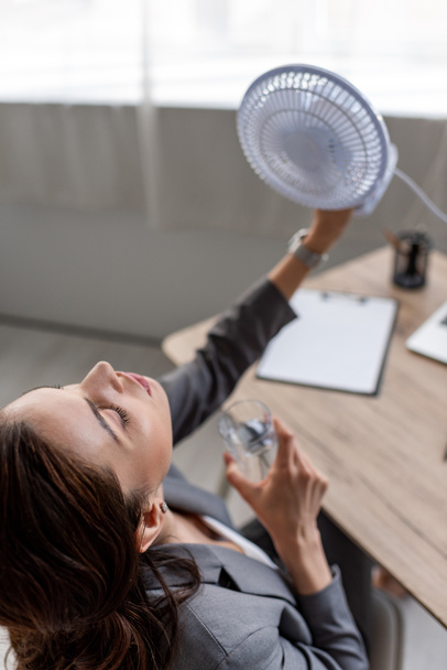 overhead view of pretty businesswoman holding blowing electric fan and glass of water while suffering from heat in office - 写真・画像