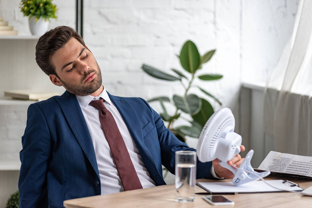 handsome exhausted businessman suffering from heat while sitting at workplace in front of blowing electric fan  - Photo, Image
