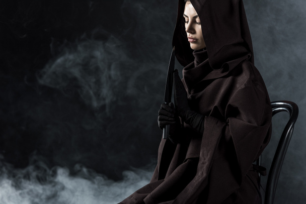 mujer en traje de la muerte sosteniendo cuchillo y sentado en silla en negro
 - Foto, imagen