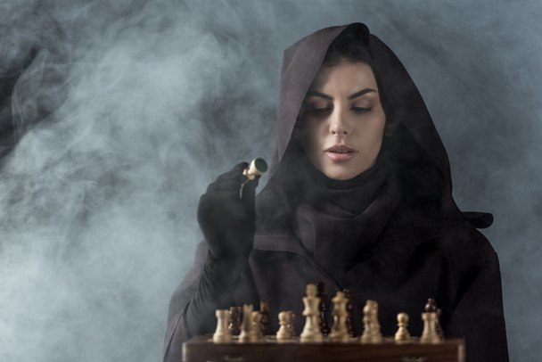 woman in death costume playing chess in smoke on black - Fotoğraf, Görsel
